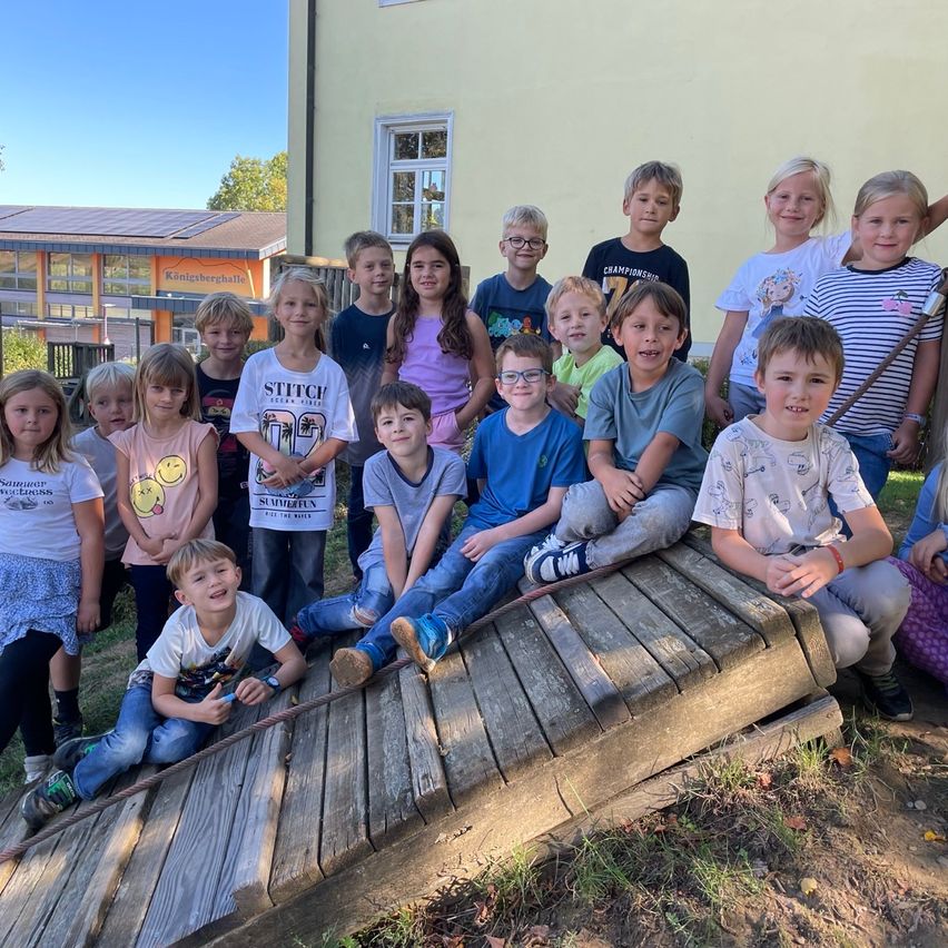 Eine Gruppe von Kindern in Freizeitkleidung posiert für ein Foto vor einem Gebäude. Einige sitzen auf einer Holzplanke, während andere hinter ihnen stehen.