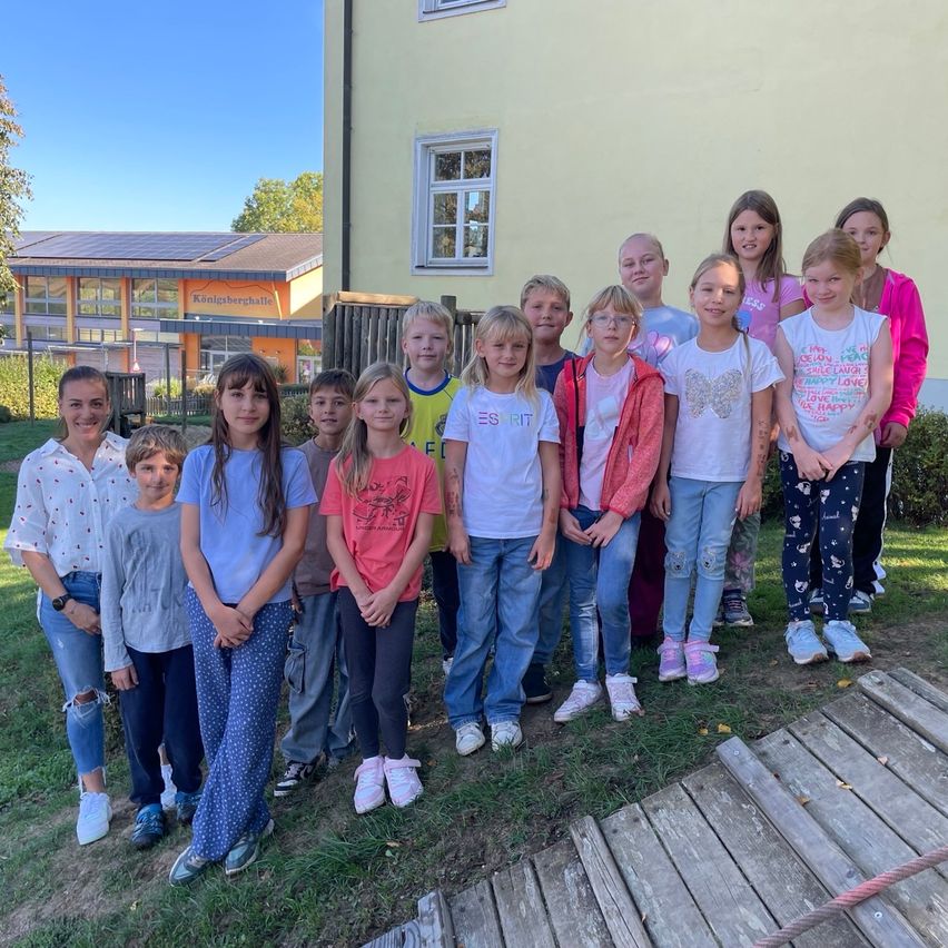 Eine Gruppe von Kindern und zwei Erwachsenen posiert für ein Foto vor einem Gebäude. Die Kinder tragen Freizeitkleidung und einige haben Armbänder. Das Gebäude hat Glasfenster und ein Dach mit Solarmodulen.