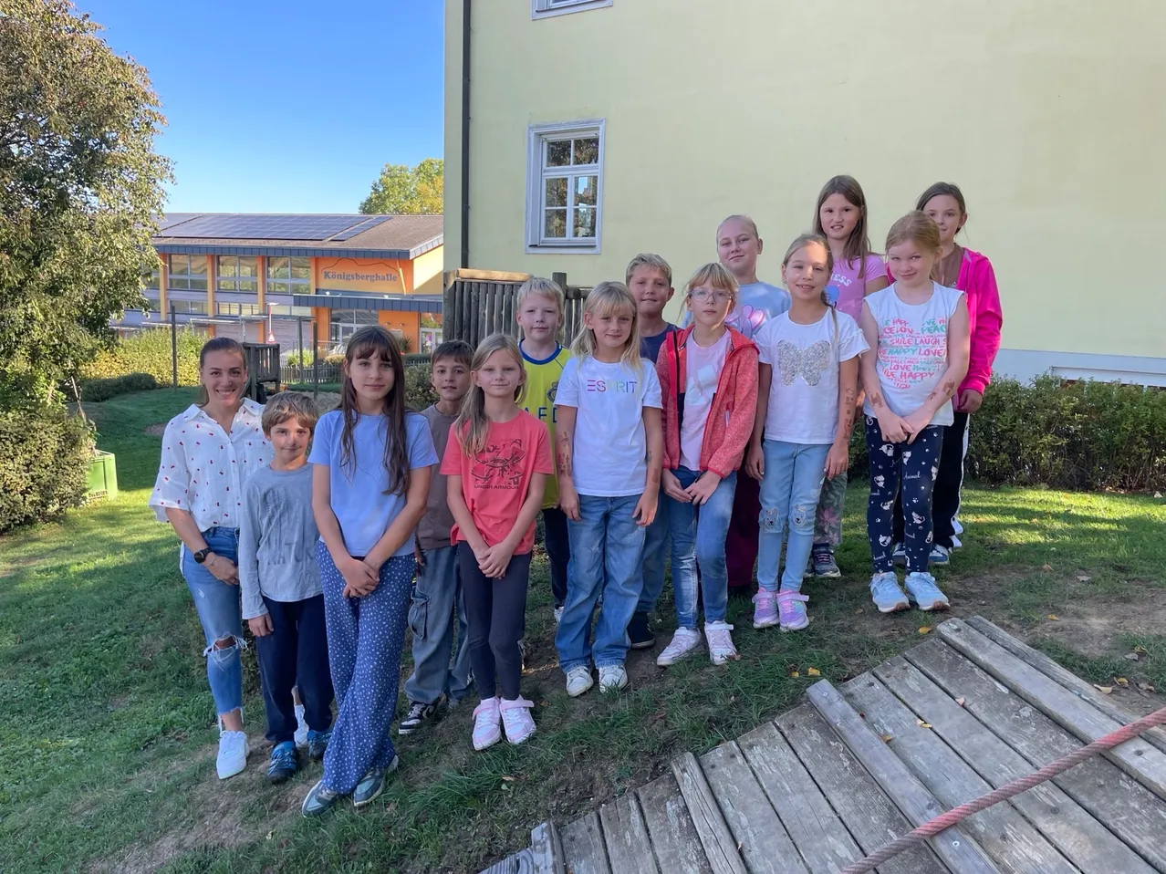 Eine Gruppe von Kindern und zwei Erwachsenen posiert für ein Foto vor einem Gebäude. Die Kinder tragen Freizeitkleidung und einige haben Armbänder. Das Gebäude hat Glasfenster und ein Dach mit Solarmodulen.