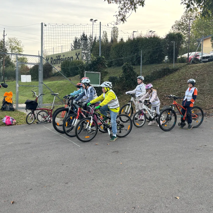 Eine Gruppe von Kindern fährt auf einem gepflasterten Bereich mit Fahrrädern. Sie tragen Helme und einige tragen reflektierende Westen.