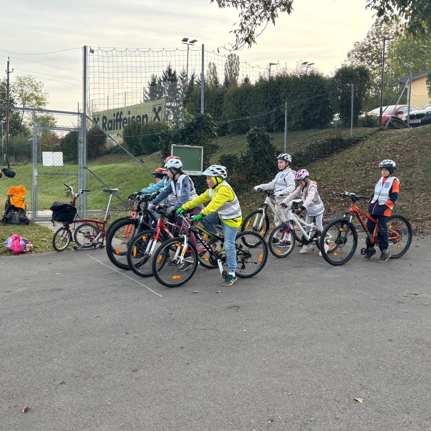Eine Gruppe von Kindern fährt auf einem gepflasterten Bereich mit Fahrrädern. Sie tragen Helme und einige tragen reflektierende Westen.