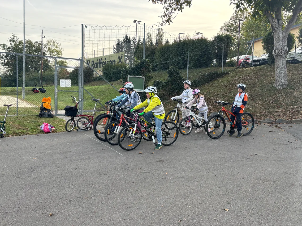 Eine Gruppe von Kindern fährt auf einem gepflasterten Bereich mit Fahrrädern. Sie tragen Helme und einige tragen reflektierende Westen.