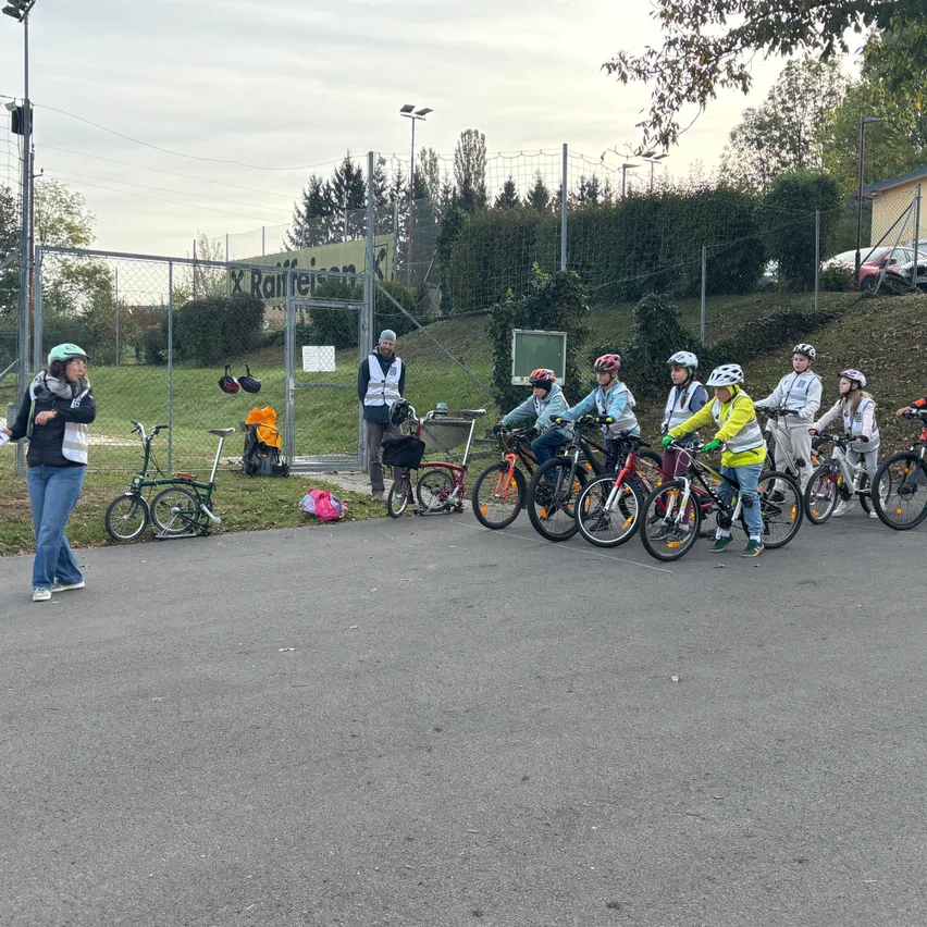 Eine Gruppe von Kindern mit Helmen steht mit ihren Fahrrädern auf einer gepflasterten Straße. Eine Frau steht in der Nähe und hält ein Stück Papier.