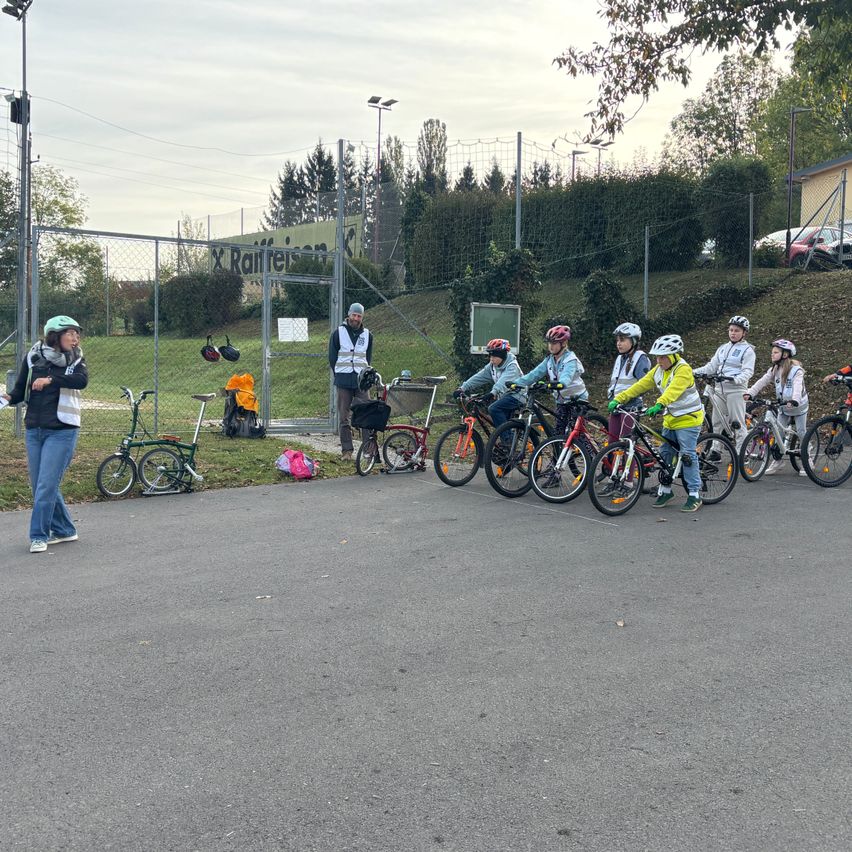 Eine Gruppe von Kindern mit Helmen steht mit ihren Fahrrädern auf einer gepflasterten Straße. Eine Frau steht in der Nähe und hält ein Stück Papier.