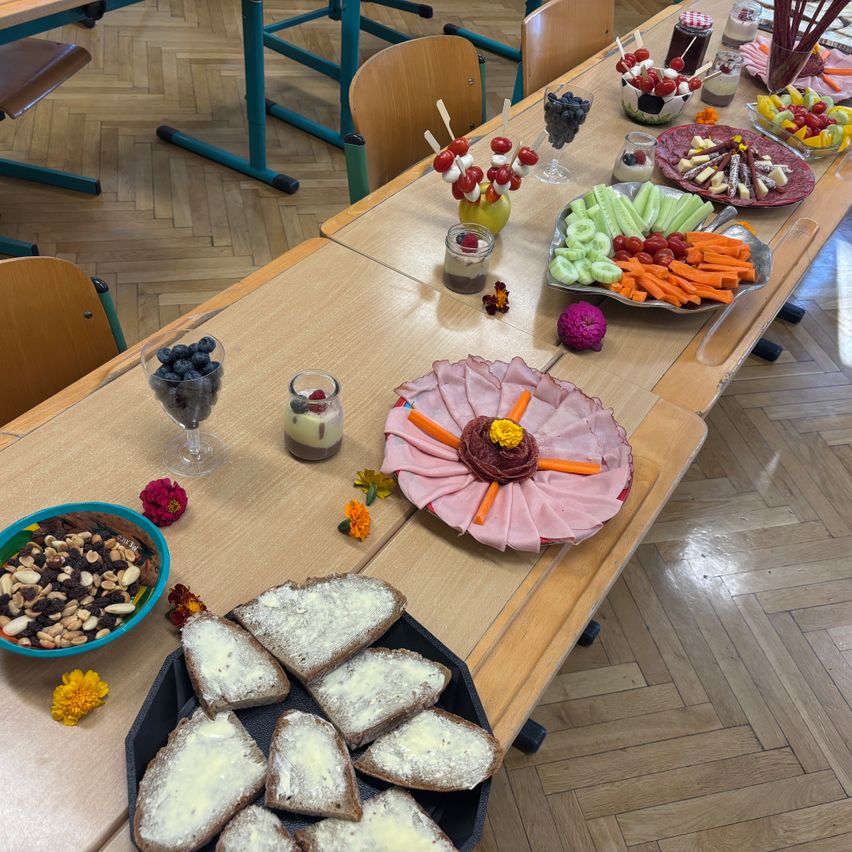 Ein langer Tisch ist mit verschiedenen Lebensmitteln wie Obst, Gemüse und Brot gedeckt. Auf dem Tisch stehen Blumen und Kerzen.