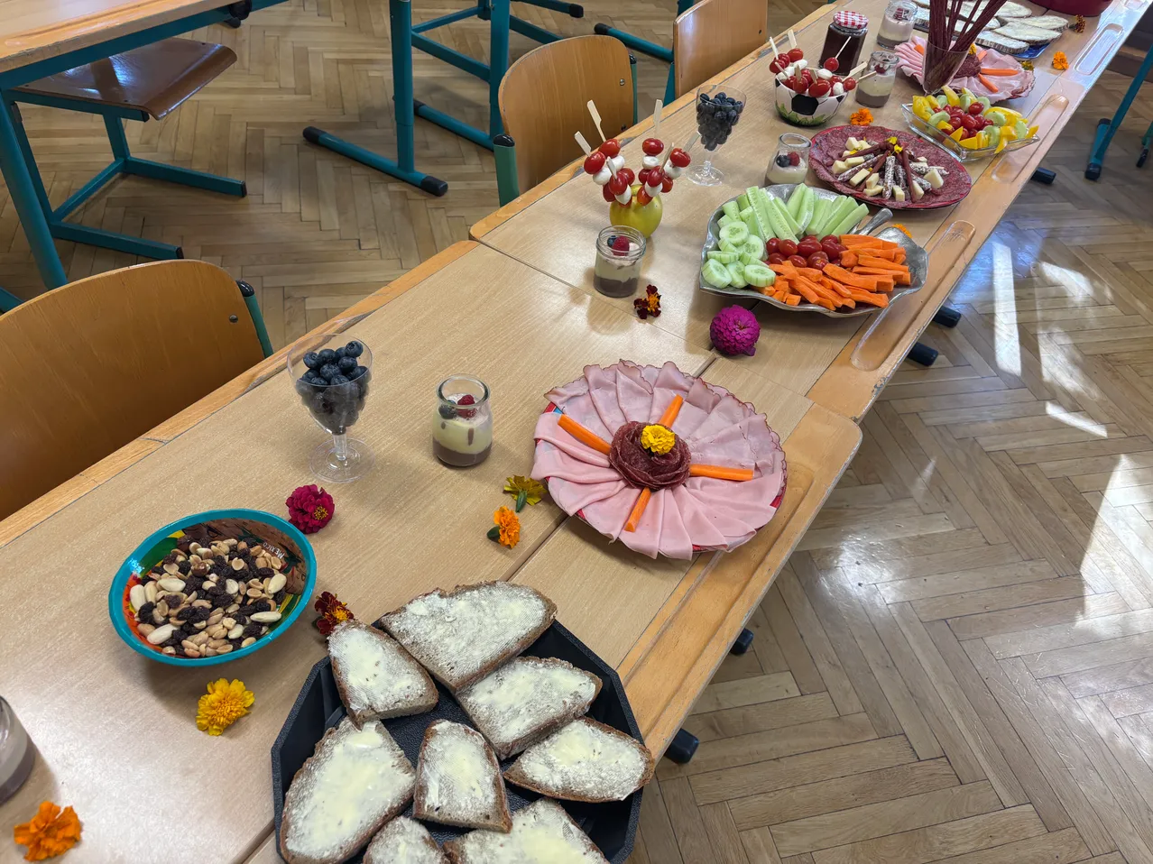 Ein langer Tisch ist mit verschiedenen Lebensmitteln wie Obst, Gemüse und Brot gedeckt. Auf dem Tisch stehen Blumen und Kerzen.