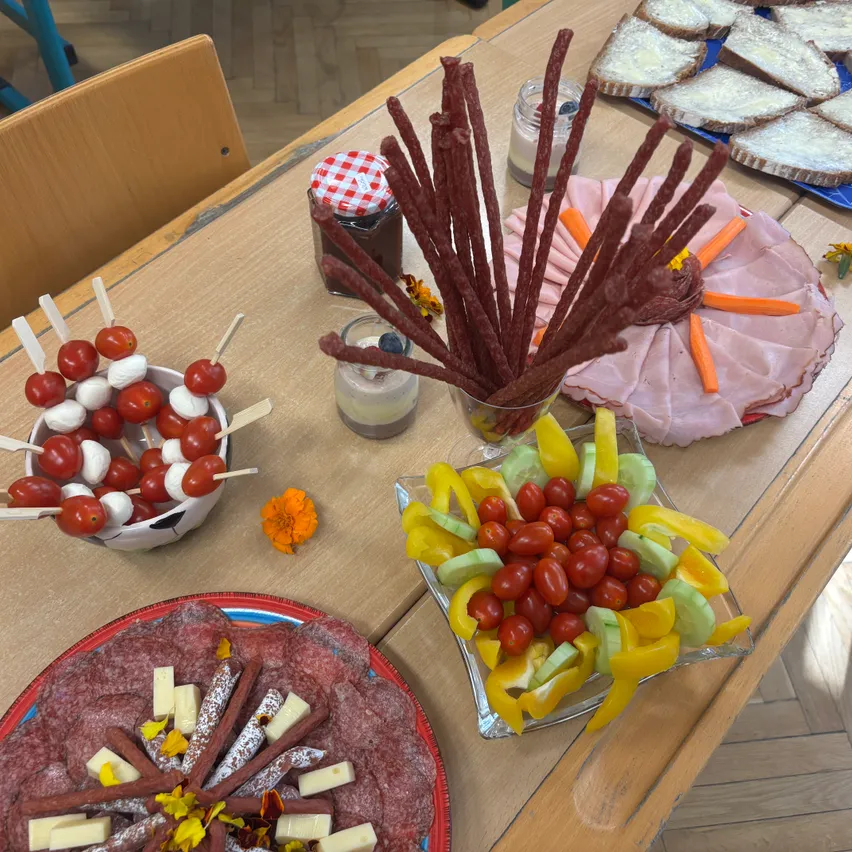 Ein Holztisch mit verschiedenen Lebensmitteln darauf. Es gibt zwei Teller mit Fleisch, einer mit einer Blumendekoration und ein Teller mit einer Mischung aus Gemüse und Früchten. Es gibt auch zwei Gläser mit Deckeln, eines mit einem rot-weiß karierten Deckel.