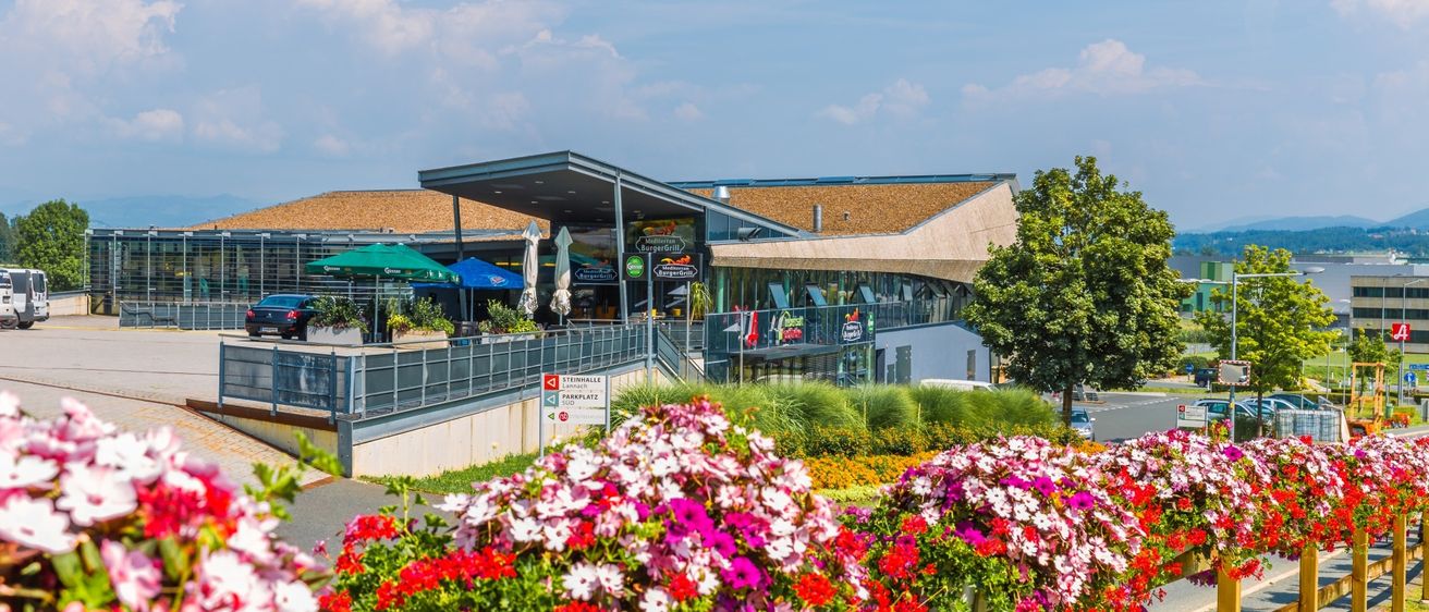 Ein modernes Gebäude mit einem gebogenen Dach beherbergt ein Restaurant, umgeben von leuchtenden Blumen und Grün, unter einem klaren blauen Himmel.
