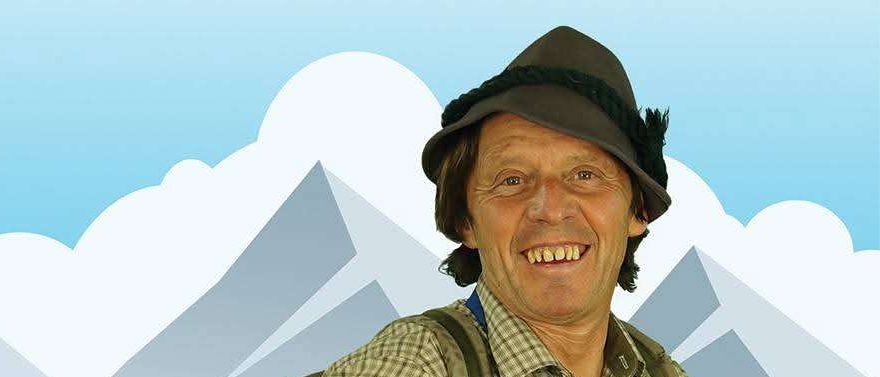 Ein Mann in Hut und Schürze lächelt in die Kamera, trägt einen Rucksack in einem grünen Feld mit Bergen und blauem Himmel.