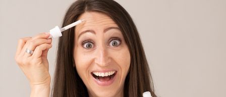 Eine Frau hält eine Flasche Serum und trägt es auf ihre Stirn auf, sie lächelt breit.