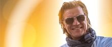 Ein Mann mit lächelndem Gesicht steht mit verschränkten Armen, trägt eine Sonnenbrille und einen Schal vor einem hellen gelben Hintergrund.