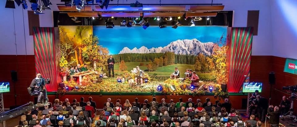 Eine Bühnenaufführung mit einer landschaftlichen Kulisse von Bergen, Menschen sitzen im Publikum, einige in traditioneller Kleidung. Eine Person steht auf der Bühne, wahrscheinlich bei einer Aufführung.
