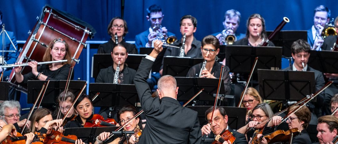 Ein Symphonieorchester spielt auf der Bühne, mit Musikern, die verschiedene Instrumente spielen. Der Dirigent führt mit erhobener Hand. Das Publikum sitzt vor ihnen.
