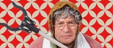 Eine ältere Frau mit Schal und Brille, die ein langes Werkzeug hält, steht vor einem rot-weiß gemusterten Hintergrund.