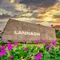 Ein großes Steinschild mit dem Wort Lannach in weißen Buchstaben steht vor einem Garten mit lila Blumen und einem bewölkten Himmel.