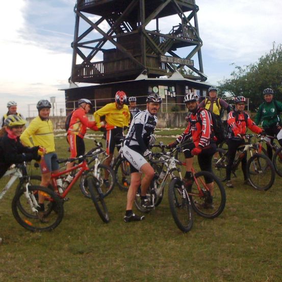 Eine Gruppe von Radfahrern mit Helmen und Fahrradausrüstung posiert vor einem Turm und einem Gebäude auf einem Grasfeld.