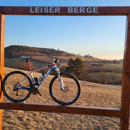 Ein Fahrrad ist vor einem Schild mit der Aufschrift 'LEISER BERGE' ausgestellt. Es befindet sich auf einem grasbewachsenen Hügel mit einem Turm in der Ferne unter einem klaren blauen Himmel.
