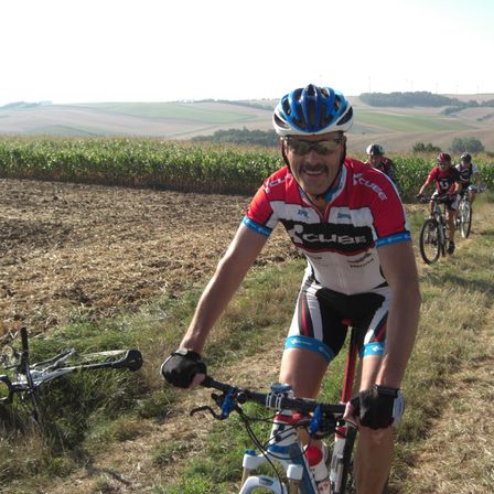 Ein Mann in einem Radtrikot fährt ein Fahrrad auf einem Feld. Hinter ihm fahren andere Radfahrer. Der Hintergrund zeigt ein Maisfeld und Hügel.