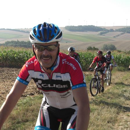 Ein Mann in einem Radtrikot mit einem blau-weißen Helm fährt ein Fahrrad auf einem Grasfeld. Hinter ihm fahren zwei Radfahrer in der Ferne. Das Feld ist von einem Maisfeld umgeben.