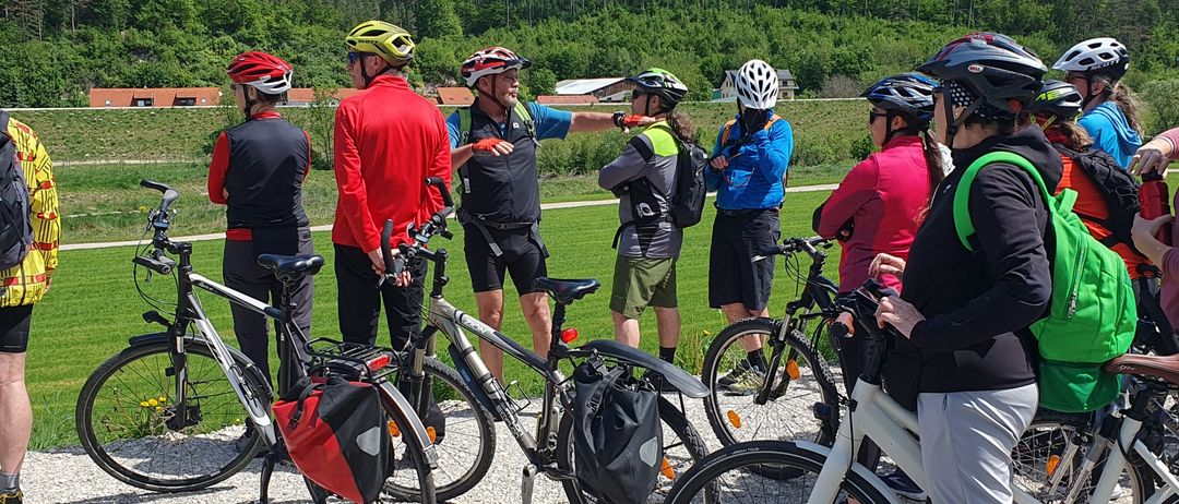 Eine Gruppe von Radfahrern versammelt sich auf Schotter, mit Fahrrädern und Helmen. Im Hintergrund befindet sich ein Hügel mit Grünflächen und einem Haus.