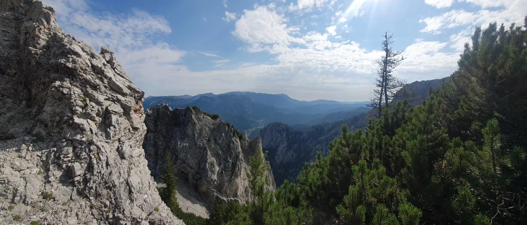 Eine Berglandschaft mit felsigen Klippen, sonnenbeschienenem Himmel und üppigem grünem Baumbestand. Der Blick ist von einem hohen Standpunkt aus.