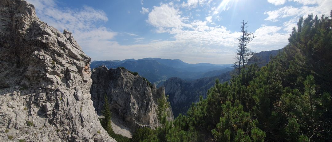 Eine Berglandschaft mit felsigen Klippen, sonnenbeschienenem Himmel und üppigem grünem Baumbestand. Der Blick ist von einem hohen Standpunkt aus.