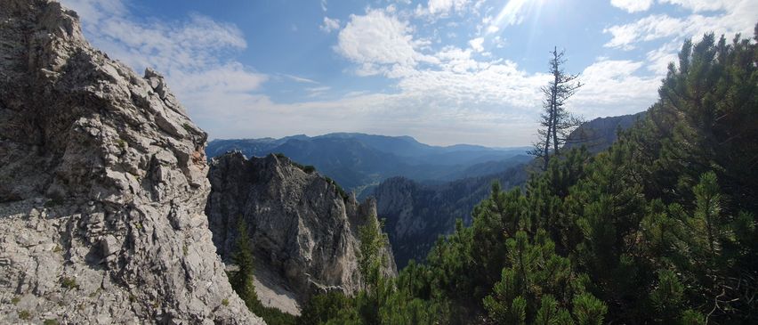 Eine Berglandschaft mit felsigen Klippen, sonnenbeschienenem Himmel und üppigem grünem Baumbestand. Der Blick ist von einem hohen Standpunkt aus.