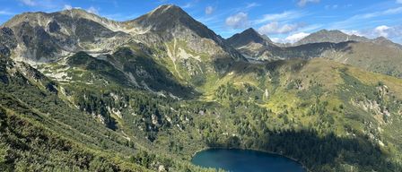 Eine Bergkette mit einem klaren See an ihrer Basis. Der See ist blau mit einer Reflexion des blauen Himmels. Die Berge sind mit Bäumen bedeckt und haben einige Felsen.