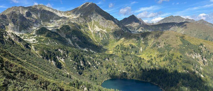 Eine Bergkette mit einem klaren See an ihrer Basis. Der See ist blau mit einer Reflexion des blauen Himmels. Die Berge sind mit Bäumen bedeckt und haben einige Felsen.
