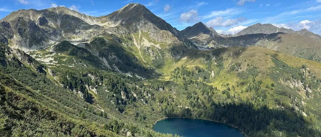 Eine Bergkette mit einem klaren See an ihrer Basis. Der See ist blau mit einer Reflexion des blauen Himmels. Die Berge sind mit Bäumen bedeckt und haben einige Felsen.