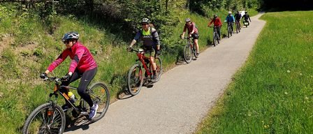 Eine Gruppe von Radfahrern fährt auf einem asphaltierten Weg, umgeben von üppigem Grün und Bäumen, unter einem klaren blauen Himmel.