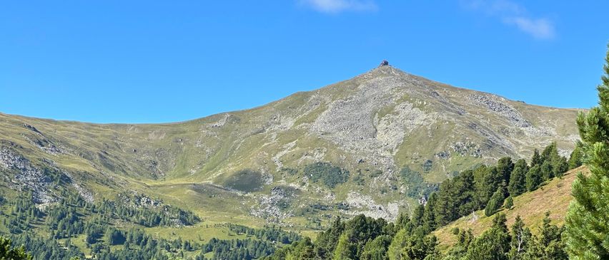 Ein Berggipfel mit einer kleinen Struktur auf der Spitze, umgeben von grüner Vegetation unter einem klaren blauen Himmel.