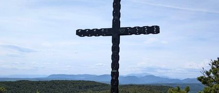 Ein großer, verzierter schwarzer Kreuz steht auf einem Hügel, umgeben von üppigem Grün und einem klaren blauen Himmel.