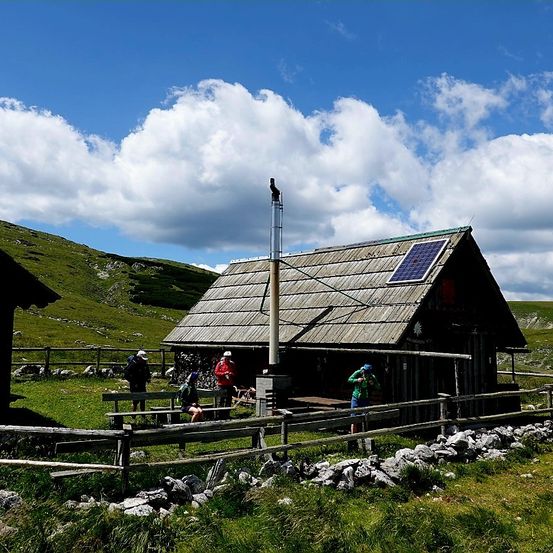 Ein kleines Holzhaus mit einem Solarpanel auf dem Dach, umgeben von einem Zaun und grünem Gras. Mehrere Menschen stehen davor.