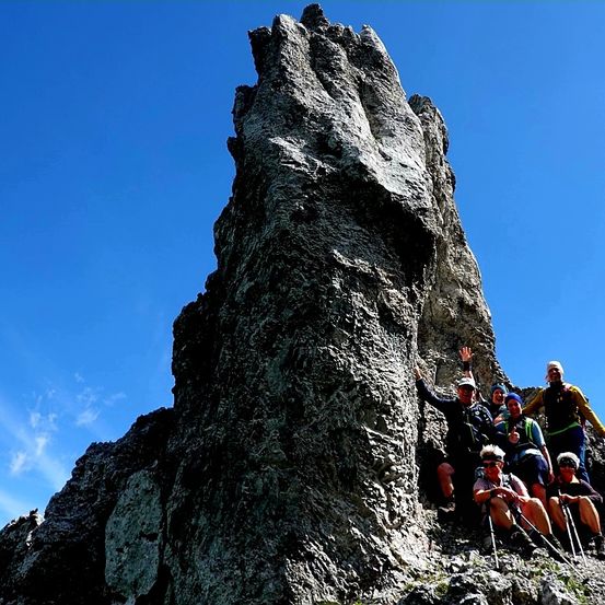 Eine Gruppe von Wanderern posiert für ein Foto auf einem felsigen Berg unter einem klaren blauen Himmel. Einige sitzen auf Felsen, während andere stehen.