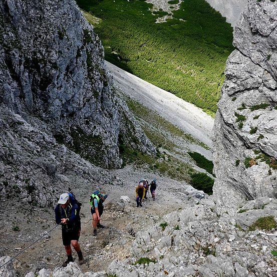 Eine Gruppe von Wanderern erklimmt einen steilen, felsigen Pfad mit grünem Gras an den Seiten. Der Weg ist eng und schwierig.