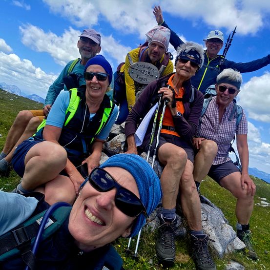 Eine Gruppe von Wanderern sitzt auf einem grasbewachsenen Berghang und lächelt für ein Foto. Sie tragen Hüte, Brillen und Rucksäcke, einer hält ein Holzschild mit 'Steboden' und '4839m'.