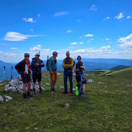Fünf Wanderer stehen auf einem grasigen Berggipfel unter einem blauen Himmel mit Wolken und halten Trekkingstöcke. Sie tragen Rucksäcke und Mützen. Dahinter ist ein Blick auf Berge und Täler.