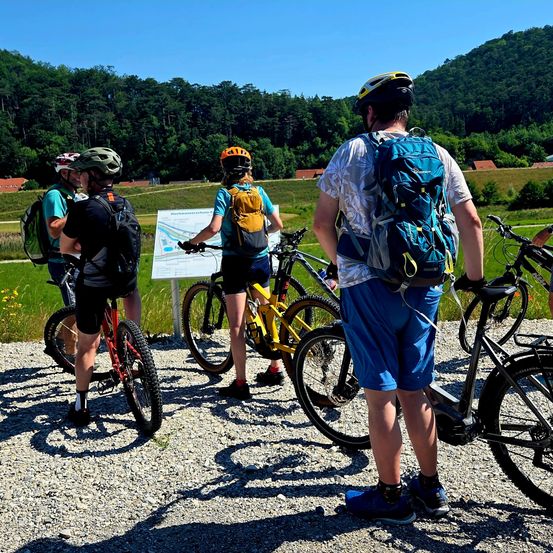 Eine Gruppe von Radfahrern bereitet sich auf eine Fahrt in einer malerischen Gegend mit Bäumen und einem Hügel im Hintergrund vor.