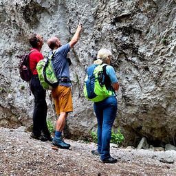 Drei Personen stehen auf einem felsigen Pfad und tragen Rucksäcke. Ein Mann zeigt auf die Wand.
