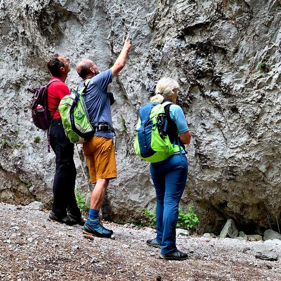 Drei Personen stehen auf einem felsigen Pfad und tragen Rucksäcke. Ein Mann zeigt auf die Wand.