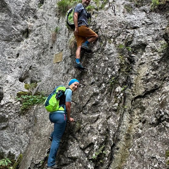 Zwei Menschen klettern einen felsigen Abhang hinauf. Einer trägt einen grünen Rucksack und blaue Jeans, der andere einen blauen Rucksack und orangefarbene Shorts. Beide lächeln und sehen glücklich aus.