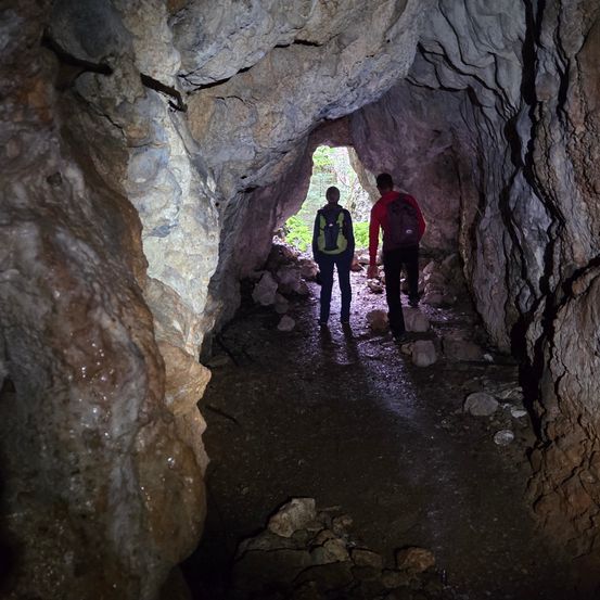 Zwei Personen mit Rucksäcken gehen durch eine enge Höhle, beleuchtet durch Licht von draußen.