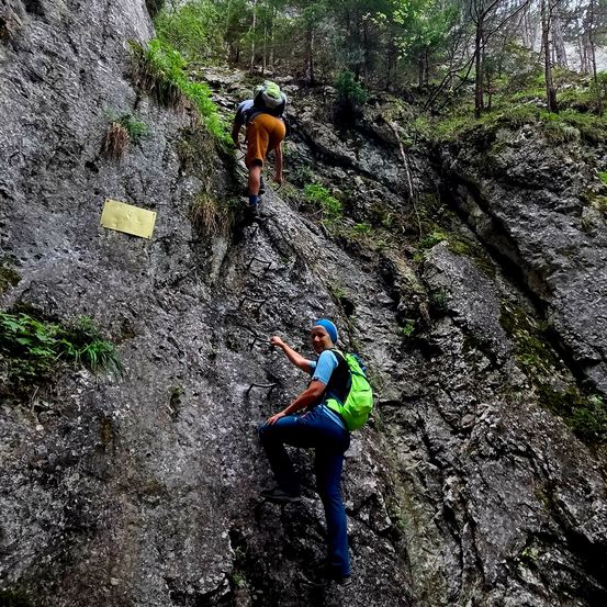 Zwei Wanderer klettern einen felsigen Hang hinauf. Einer trägt einen grünen Rucksack und der andere orangefarbene Shorts.