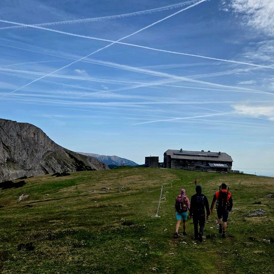 Drei Wanderer gehen auf einem grasbewachsenen Bergpfad unter einem blauen Himmel mit Flugzeugkondensstreifen. Ein kleines Gebäude ist in der Ferne zu sehen.