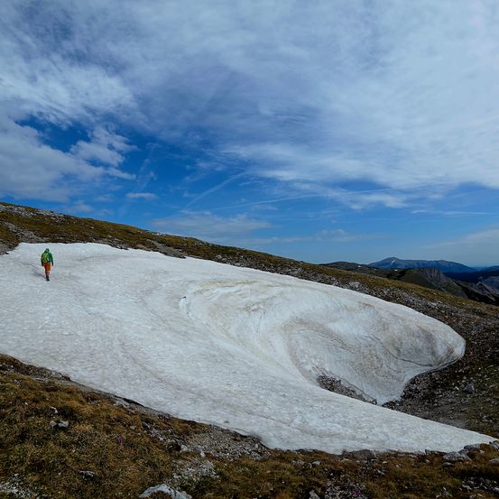 Eine Person in einer grünen Jacke läuft auf einem schneebedeckten Pfad auf einem Berg unter einem blauen Himmel mit verstreuten Wolken.