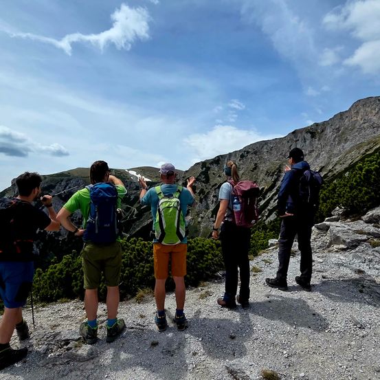 Fünf Wanderer stehen auf einem felsigen Pfad und blicken auf eine Bergkette, mit dem Rücken zur Kamera. Der Himmel ist blau mit verstreuten Wolken.