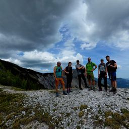 Sechs Personen stehen auf einem felsigen Berggipfel und posieren für ein Foto unter einem teilweise bewölkten Himmel.