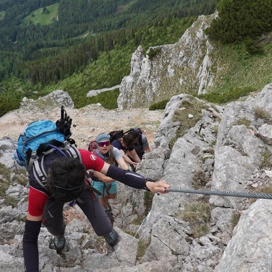 Drei Wanderer klettern einen felsigen Pfad in einem Berggebiet hinauf, einer hält ein Seil. Sie sind mit Rucksäcken und Sonnenbrillen ausgestattet.