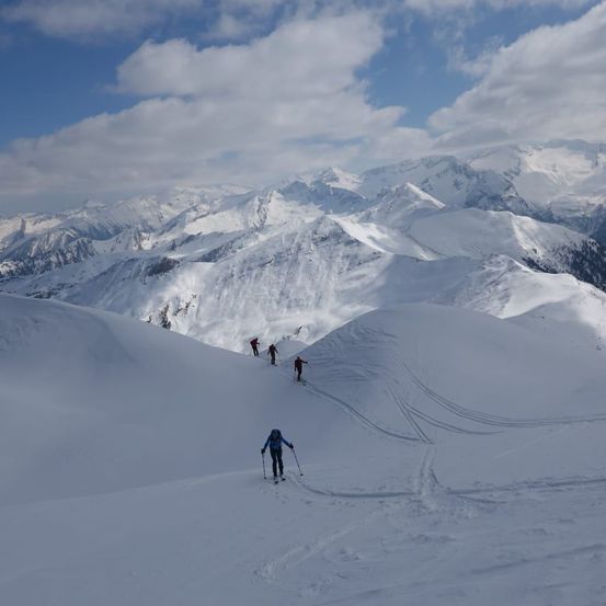 Fünf Skifahrer bewegen sich durch eine verschneite Berglandschaft unter einem blauen Himmel mit verstreuten Wolken. Sie sind mit Skistöcken und Rucksäcken ausgestattet.